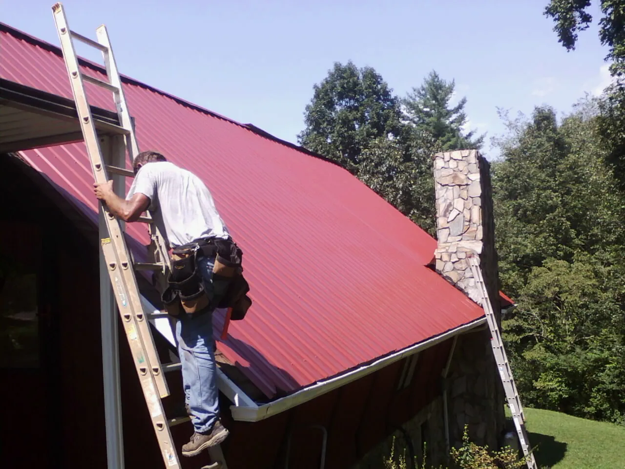 Roofer on a ladder beside a red metal roof and stone chimney — Soffit & Fascia Repair by Roofing Brothers in Naytahwaush