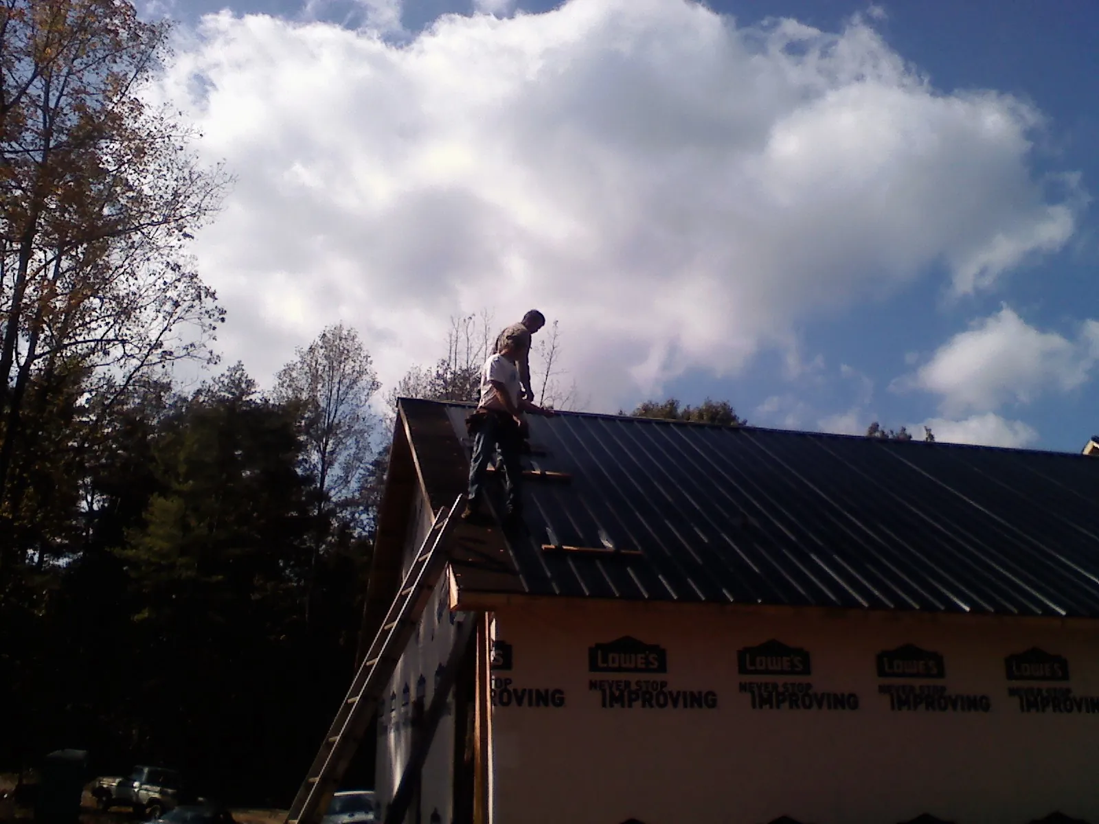 Roofer fastening dark metal panels high against a cloudy sky, from a Roof Inspection job in Naytahwaush